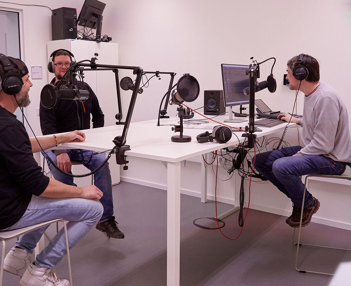 Drei Personen mit Kopfhörern nehmen in einem hellen Podcast-Studio auf
